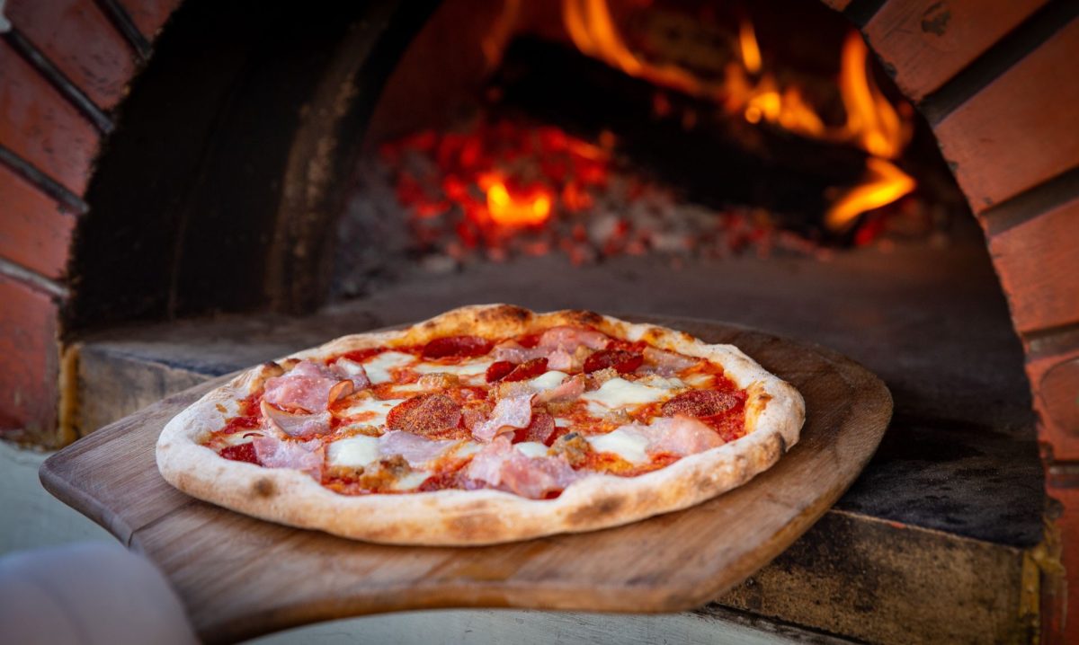 Pizza with pepperoni and ham slices on melted cheese, resting on a wooden paddle in front of a brick oven with glowing embers.