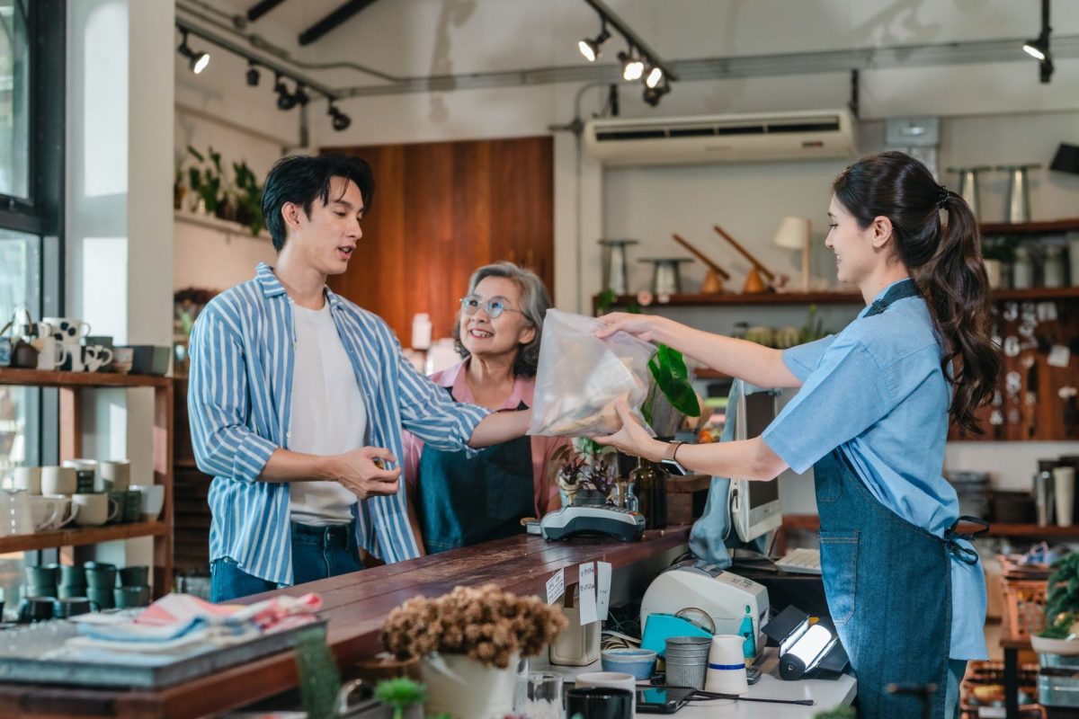 Shopkeeper hands a bag to a customer at a wooden counter, with an older smiling woman in the background.